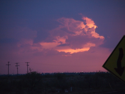 [A view of the distant horizon which has three power poles on the left and a multitude of lights at the juncture of the land and sky. The sky is shades of violet and blue with one large cloud way above the land. The cloud color ranges from white to shades of light purple and blue.]